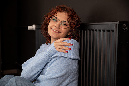 笑着的女孩坐在热散热器旁边 靠着她倾斜生态房间回暖持有暖气眼镜活力系统安装热能图片
