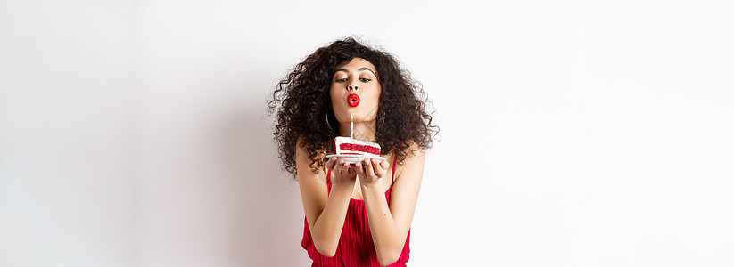 喜剧节和庆祝概念 穿红礼服的浪漫女郎庆祝生日 在一天蛋糕上吹蜡烛并许愿 站在白色背景之上发型嘴唇工作室促销投标庆典假期黑发情感微图片