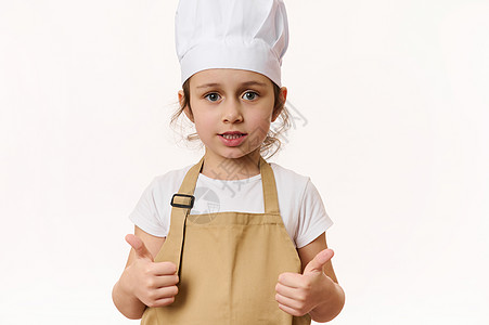 戴着厨师帽和米色围裙的白人可爱小女孩竖起大拇指 看着白色背景中的相机图片