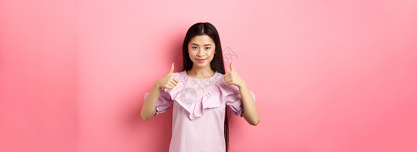 好样的 笑得漂亮 微笑着的亚洲女孩穿著洋装露出拇指 像和批准产品 赞美很多 站在粉红背景上投标购物女性学生女士工作室化妆品青少年图片