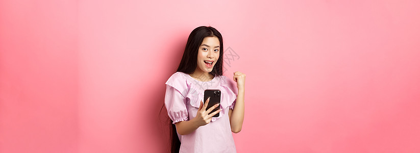 网上购物 满意的亚洲女孩在手机上获胜 说是 握拳 手持智能手机 站在粉红色背景下学生上网青少年互联网女性化妆品技术电话女士购物图片