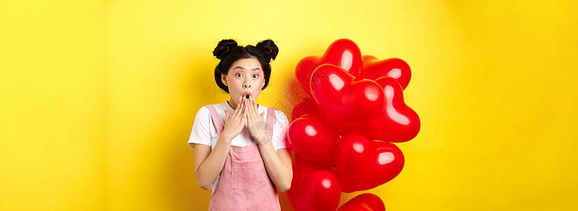 令人印象深刻的韩国女孩说 哇 在张开的嘴边喘气和握手 盯着摄影机大吃一惊 站在浪漫的红心气球旁 黄色背景促销伙伴假期微笑情人女士图片