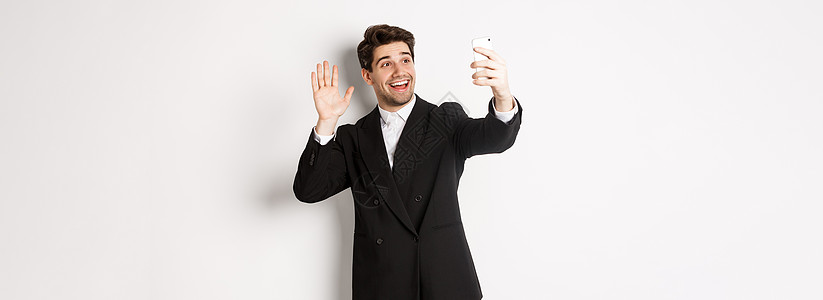 穿着西装的英俊男子的形象 在智能手机摄像头前进行视频通话和挥手 录制视频 问候某人 站在白色背景下广告房地产男人成功互联网代理人图片