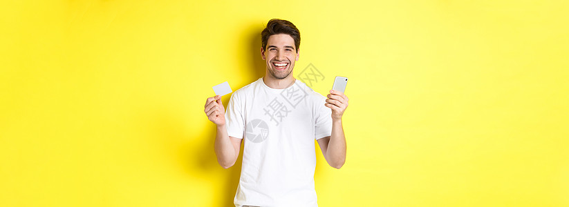 快乐的年轻人在网上用智能手机购物 持有信用卡和微笑 站在黄色背景上站立着电话情绪闲暇促销商业男性技术工具广告互联网图片