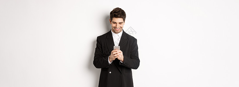 穿着黑西装的英俊时尚商务人士的肖像 写一个信息 微笑和看着智能手机 站在白色背景上房地产雇主购物员工横幅工具广告技术互联网男人图片