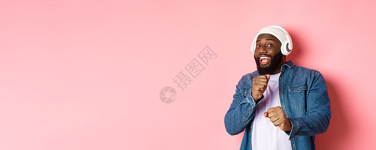 欢乐的非洲美国人跳舞 用耳机听音乐和看摄影机 站在粉红背景上促销横幅男人手机电话快乐情感技术学生衬衫图片