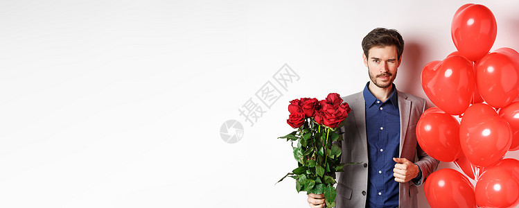 自信的男友穿着西装去浪漫约会 对着镜头眨眼 拿着一束红玫瑰 站在心形气球附近眨眼 站在白色背景上展示男性花朵玫瑰纪念日感情花束男图片
