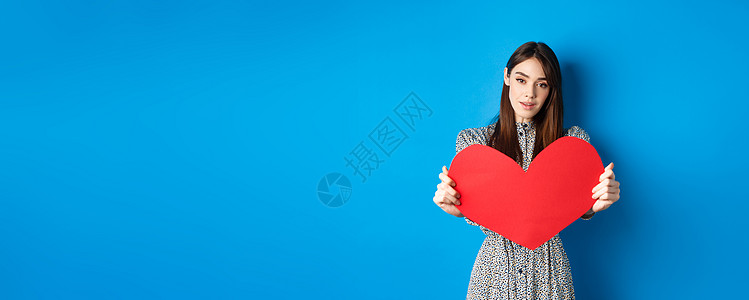 情人节和关系概念 穿着礼服的年轻女求婚者伸出双手 给你红色的大心脏 做忏悔 站在蓝色背景上浪漫地站立周年假期女孩工作室投标情感女图片