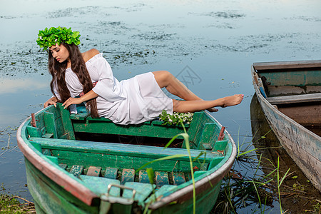 深色头发长发的女神 穿着白色古董礼服 坐在河中央的一条船上神话裙子冒充童话美人鱼魔法魅力若虫花朵女孩图片