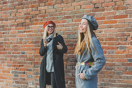 女孩在城市街道砖墙前拍摄她的女性朋友照片摄影师和青年城市生活方式概念校对 Portnoy潮人冒充摄影旅游个性青年享受生活微笑女士图片