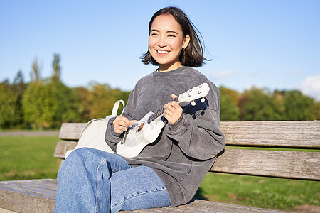 快乐的可爱女孩独自坐在公园的长椅上 弹四轮吉他 在户外享受阳光明媚的一天女性唱歌喜悦花园爱好弦琴乐器乐趣黑发幸福图片