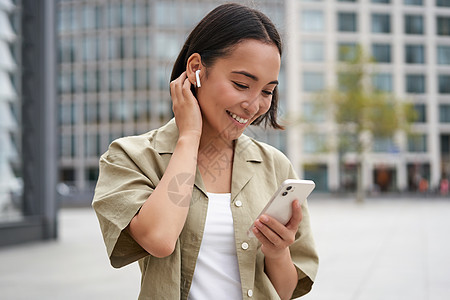 微笑的亚洲女孩用无线耳机听音乐 看着她的手机 选择音乐或播客 打电话给某人的少妇 使用耳机快乐城市出租车黑发街道互联网技术青少年图片