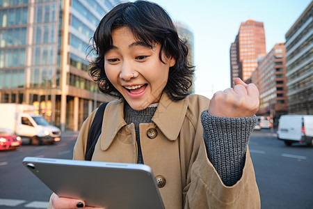 热情的亚洲女孩带着数字平板电脑 微笑着 看起来很惊讶 对一些好东西很满意 拿着数字平板电脑 站在街上学生设计师界面旅游街道艺术艺图片