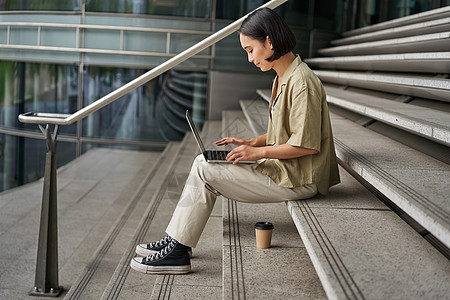 带笔记本电脑的亚洲年轻女性的侧面肖像 女学生坐在大楼外的楼梯上 在电脑上打字 喝外卖咖啡女孩视频技术女士互联网电话商业青少年城市图片
