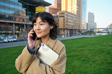 时尚的微笑女孩 穿着风衣的大学生 拿着平板电脑 用手机说话 通过电话交谈 看起来很放松 站在街上用户城市女士设计师细胞自由职业者图片