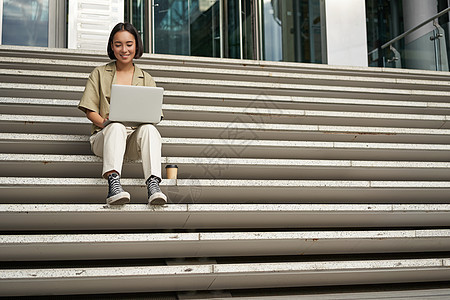 亚裔女学生坐在校园附近的楼梯上 在笔记本电脑上打字 在户外做家庭作业商业女孩视频女士电话快乐城市手机女性互联网图片