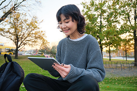 年轻微笑的韩国女孩的肖像 图形设计师 艺术家用笔工具在数字平板上画画 坐在公园中呼吸新鲜空气和抓捕 作笔记快乐互联网艺术家成人女图片