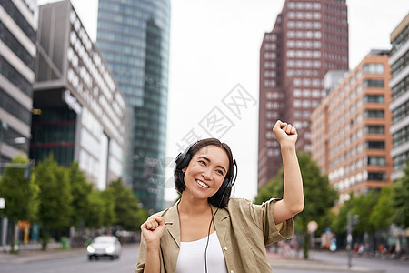 在城市聆听音乐 在摩天大楼附近街头摆姿势时 微笑着的亚洲女性跳舞 胜利 快乐和快乐的肖像青少年唱歌商业街道耳机电话技术手机喜悦黑图片