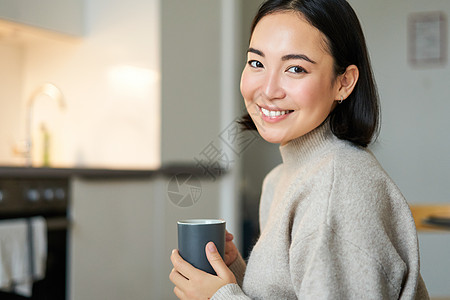 带着咖啡 喝热茶和在家舒适的笑笑的亚洲女孩的肖像 用饮料来暖和起来桌子沙发闲暇互联网女性太阳生活长椅自拍厨房图片