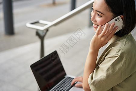 亚洲女孩的画像 学生坐在楼梯上 拿着笔记本电脑 用手机聊天 年轻女性在电脑上工作时打电话互联网视频电话城市街道技术青少年黑发商业图片