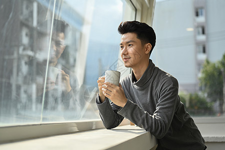 愉快的亚洲人享受着早晨咖啡 站在办公室窗户旁边 俯视城市风景图片