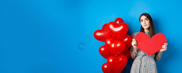 情人节 梦幻般的漂亮女士穿着裙子 拿着大红色的心形剪纸 寻找真爱 抬头沉思 站在蓝色背景的假日气球旁微笑庆典工作室女朋友礼物情感图片