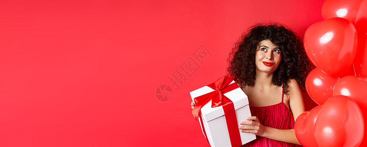 情人节和爱情观 化着浪漫妆容的漂亮女人 拿着秘密仰慕者的礼盒 梦幻般地看着左上角 站在红色背景上的心形气球盒子礼物促销惊喜纪念日图片