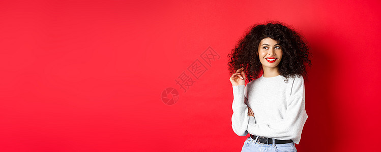 现代妇女穿着运动衫 玩卷发 仰视空旷空间 站在红色背景上站立黑发卷曲化妆品女士工作室生活促销发型女孩嘴唇图片