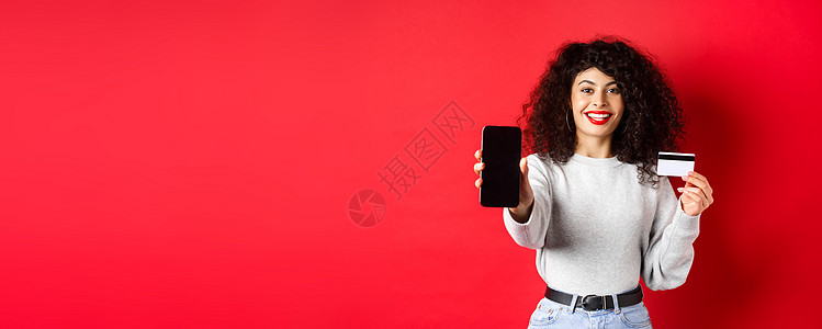展示塑料信用卡和空智能手机屏幕的快乐漂亮女孩 展示账户或应用程序 站在红色背景上电话促销生活购物广告女性卷曲工作室技术黑发图片