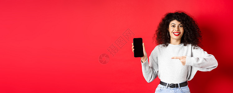 漂亮的女模特展示空的智能手机屏幕 指着手机显示屏微笑 站在红色背景下电话情感广告促销工作室购物女性黑发技术卷曲图片