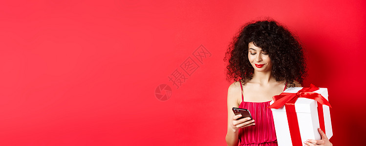手机界面情人节和购物 身穿红裙的漂亮白人女性 拿着情人的礼盒 用手机 在智能手机上阅读信息 工作室背景展示嘴唇网络电话女朋友周年纪念日盒背景