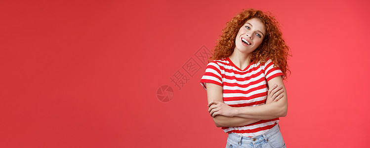 开朗无忧无虑的红发俏皮轻松的卷发女孩倾斜头微笑快乐交叉双臂胸部想要填补暑假完美假期回忆站立红色背景条纹 T 恤女士成人理发广告学图片
