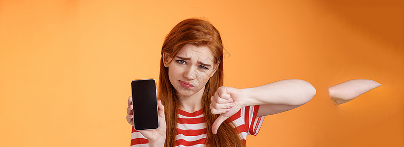 质量很差 心烦意乱的红发女顾客表情失望 显示智能手机空白显示 大拇指向下判断 不喜欢应用程序 假笑沮丧 站在心烦意乱的橙色背景互图片