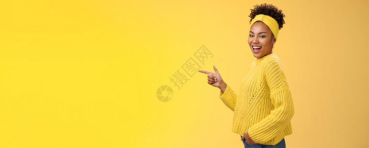 时髦可爱的女性非洲裔美国女孩 20 多岁 头带毛衣 面带微笑 面带轻浮的表情 相机转动站立的侧面 指向左侧 介绍令人敬畏的新产品图片