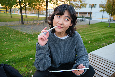 亚洲女性的画像 公园里的学生 坐在长椅上 拿着数字平板电脑和一支笔 思考 一边思考 一边做笔记 在户外做作业设计师界面药片用户成图片