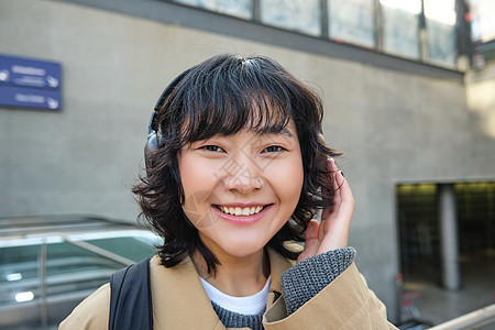 在耳机里紧贴着微笑的黑发女孩 听音乐 绕城市旅行 往返上班 站在街上互联网游客车站咖啡女士女性快乐旅游街道歌曲图片