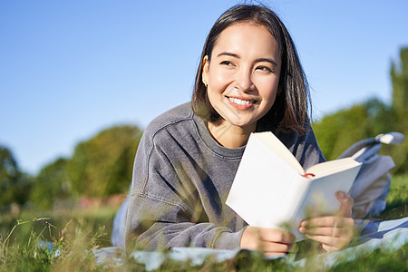 可爱的韩国女孩的画像 在公园里躺在草地上读书 手里拿着最喜欢的书放松 开心地笑着背包快乐乐趣黑发成人微笑读者幸福花园喜悦图片