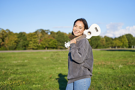 年轻的时装女孩 旅行者 抱着她的乌合乐 在公园户外玩耍和微笑歌曲唱歌乐趣吉他女性黑发弦琴喜悦花园幸福图片