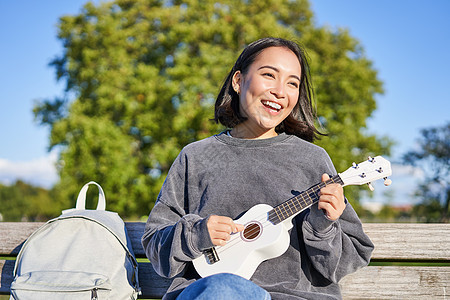 可爱的年轻女子坐在阳光明媚的公园长椅上 背着背包坐在书架上 弹四轮吉他和歌唱花园歌曲爱好微笑乐趣唱歌喜悦音乐乐器成人图片