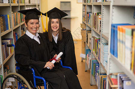 快乐的年轻女性和穿着研究生礼服坐在轮椅上的妇女 在图书馆拥有文凭 包容性教育学生教育仪式帽子微笑自由学士成就学习机动性图片