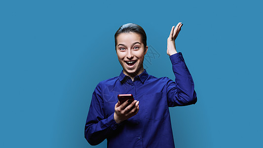 惊异的兴奋激动 震惊的年轻女性 带着智能手机 在蓝工作室背景屏幕青少年学生技术成人电话互联网蓝色女士黑发图片