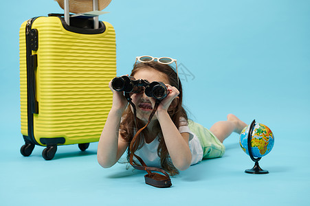 可爱的小女孩透过望远镜看望 躺在一个地球和黄色手提箱附近 背着蓝色背景;旅行概念图片