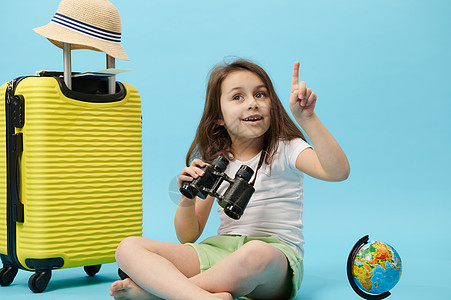 可爱的小旅行者女孩在蓝色背景的复制空间上指尖 坐在黄色手提箱和地球附近 (b)图片