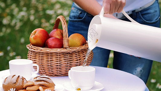 在洋甘菊草坪的背景下 女人的手将茶从一个白色的水壶里倒进一个白色的杯子 一个热水瓶 桌子上还有一个篮子 里面装着红苹果图片