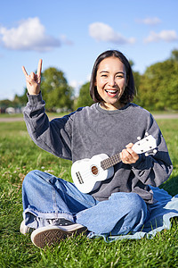 快乐的亚洲女孩在公园里玩着乐 展示摇滚 重金属角标志和微笑 在户外玩得开心女性音乐幸福歌曲成人喜悦爱好音乐家弦琴乐趣图片