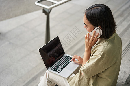 亚洲女孩的画像 学生坐在楼梯上 拿着笔记本电脑 用手机聊天 年轻女性在电脑上工作时打电话女士青少年黑发快乐街道技术城市视频商业互图片