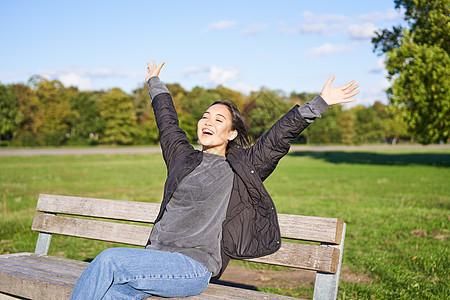 快乐的亚洲女人伸着她的手 坐在长凳上满脸兴奋 笑笑高兴 感受自由 在公园里享受一天长椅女孩成人爱好一口气幸福女性微笑乐趣黑发图片