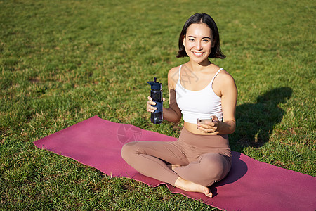 微笑着年轻的年轻女性坐在草地上 在公园做运动 修草坪 喝瓶子水工作女孩福利瑜伽快乐成人太阳训练运动装日落图片