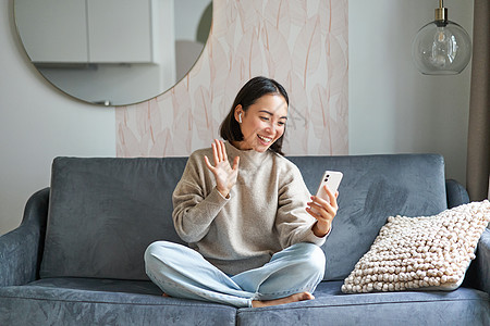 亚洲女性坐在沙发上用智能手机的肖像 在手机屏幕上挥手 在相机上挥手 视频聊天 与某人交谈房子互联网音乐女士长椅电话家具房间衬衫生图片