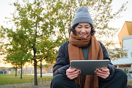 穿着暖和衣服的亚洲女孩的画像 坐在长椅上 拿着数字平板电脑和图形笔 开心地笑着 在寒冷的天气里在户外画画药片城市用户街道女士笔记图片